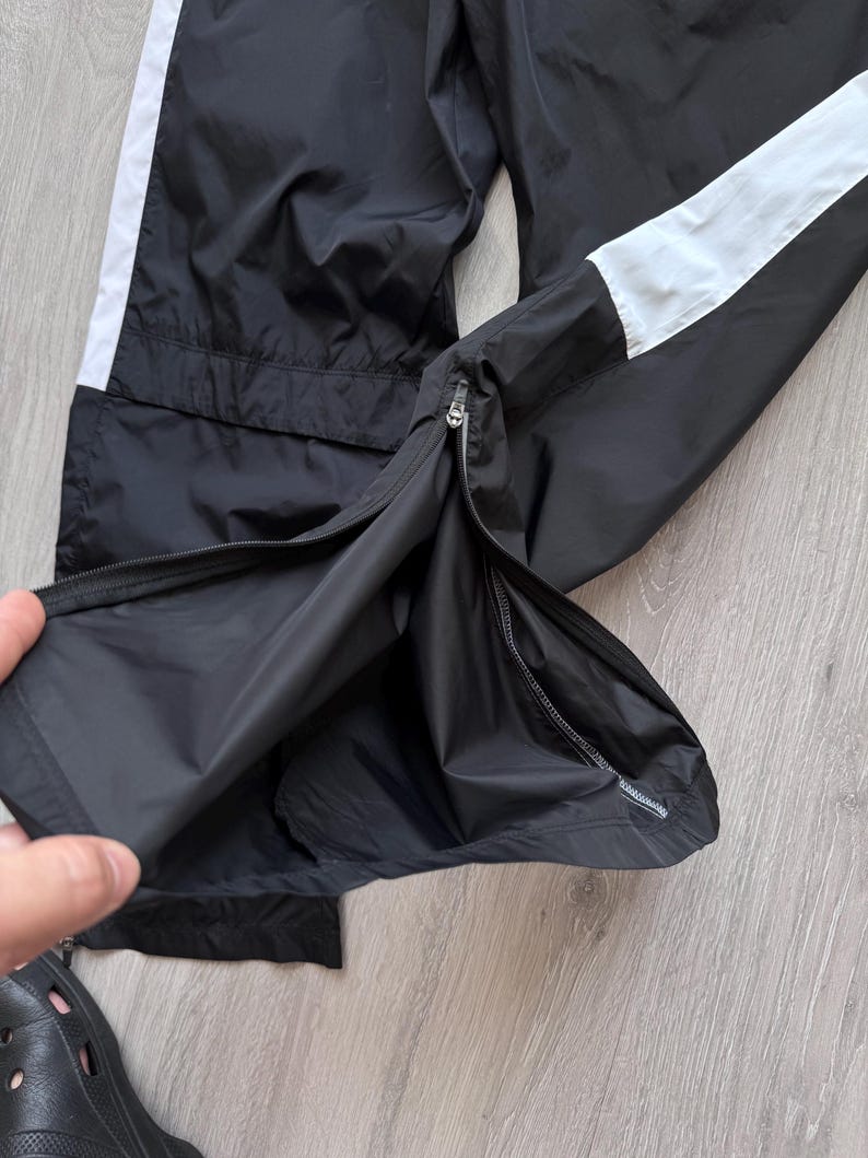 Peut inclure: Pantalon de sport noir avec une bande blanche sur le c&ocirc;t&eacute;. Le pantalon a une fermeture &eacute;clair au niveau de la cheville. Le mat&eacute;riau semble &ecirc;tre un tissu l&eacute;ger, possiblement r&eacute;sistant &agrave; l'eau.