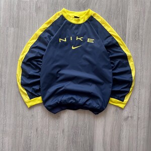 May include: Navy blue Nike pullover with yellow accents on the sleeves and collar. The Nike logo is printed in yellow across the chest. The garment is made of a lightweight material.
