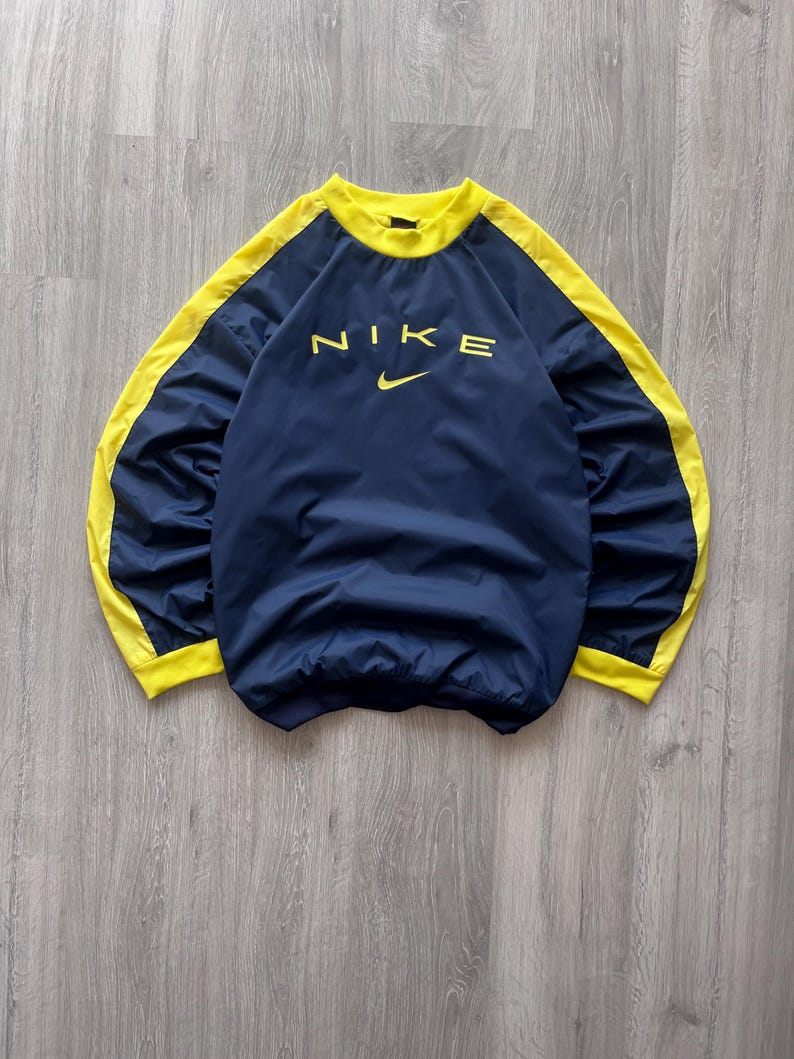 May include: Navy blue Nike pullover with yellow accents on the sleeves and collar. The Nike logo is printed in yellow across the chest. The garment is made of a lightweight material.