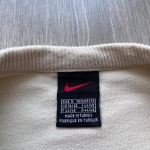Puede incluir: Primer plano de una prenda de color crema con una etiqueta negra. La etiqueta presenta un logotipo Nike rojo y texto blanco que indica la talla, la altura y el origen. El texto incluye "MADE IN TURKEY" y "FABRIQUE EN TURQUIE".