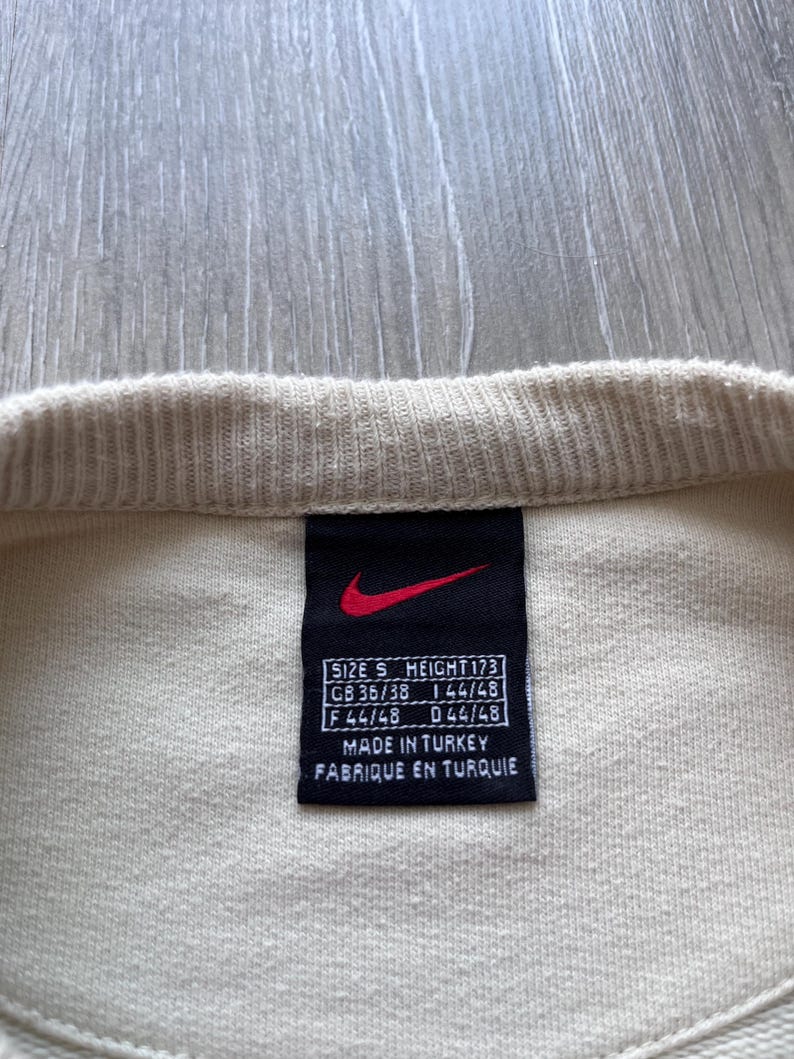 Puede incluir: Primer plano de una prenda de color crema con una etiqueta negra. La etiqueta presenta un logotipo Nike rojo y texto blanco que indica la talla, la altura y el origen. El texto incluye "MADE IN TURKEY" y "FABRIQUE EN TURQUIE".