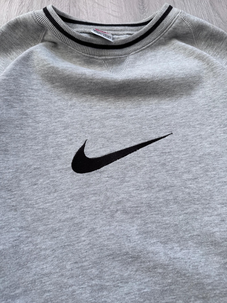 May include: A light gray sweatshirt with a black Nike swoosh logo. The crew neck and cuffs have black and white striped detailing. The sweatshirt is made of a soft, textured fabric.