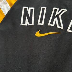 Puede incluir: Sudadera negra con la palabra "NIKE" en letras blancas. El logo de Nike es amarillo. Las mangas tienen rayas amarillas y blancas.