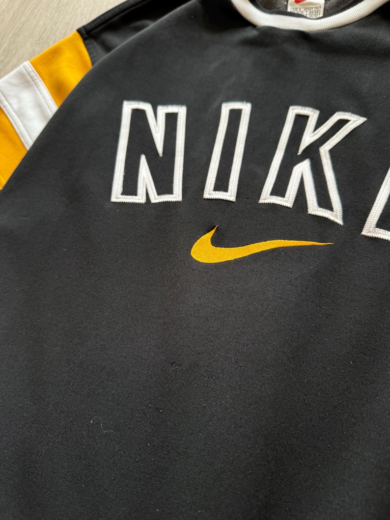Puede incluir: Sudadera negra con la palabra "NIKE" en letras blancas. El logo de Nike es amarillo. Las mangas tienen rayas amarillas y blancas.