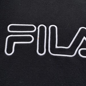 Peut inclure: Gros plan d'un sweat-shirt noir avec le mot "FILA" brod&eacute; en blanc sur la poitrine. Les lettres sont &eacute;paisses et soulign&eacute;es, cr&eacute;ant un design contrast&eacute; et audacieux. Le tissu semble doux et l&eacute;g&egrave;rement textur&eacute;.