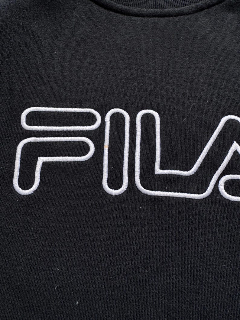 Peut inclure: Gros plan d'un sweat-shirt noir avec le mot "FILA" brod&eacute; en blanc sur la poitrine. Les lettres sont &eacute;paisses et soulign&eacute;es, cr&eacute;ant un design contrast&eacute; et audacieux. Le tissu semble doux et l&eacute;g&egrave;rement textur&eacute;.