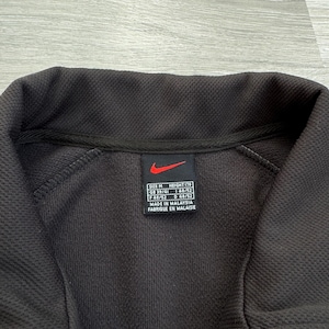 Puede incluir: Primer plano de una chaqueta deportiva negra Nike. La chaqueta tiene un patr&oacute;n texturizado y un cuello negro. Se ve una etiqueta con el logotipo de Nike e informaci&oacute;n sobre la talla. La chaqueta est&aacute; hecha en Malasia.
