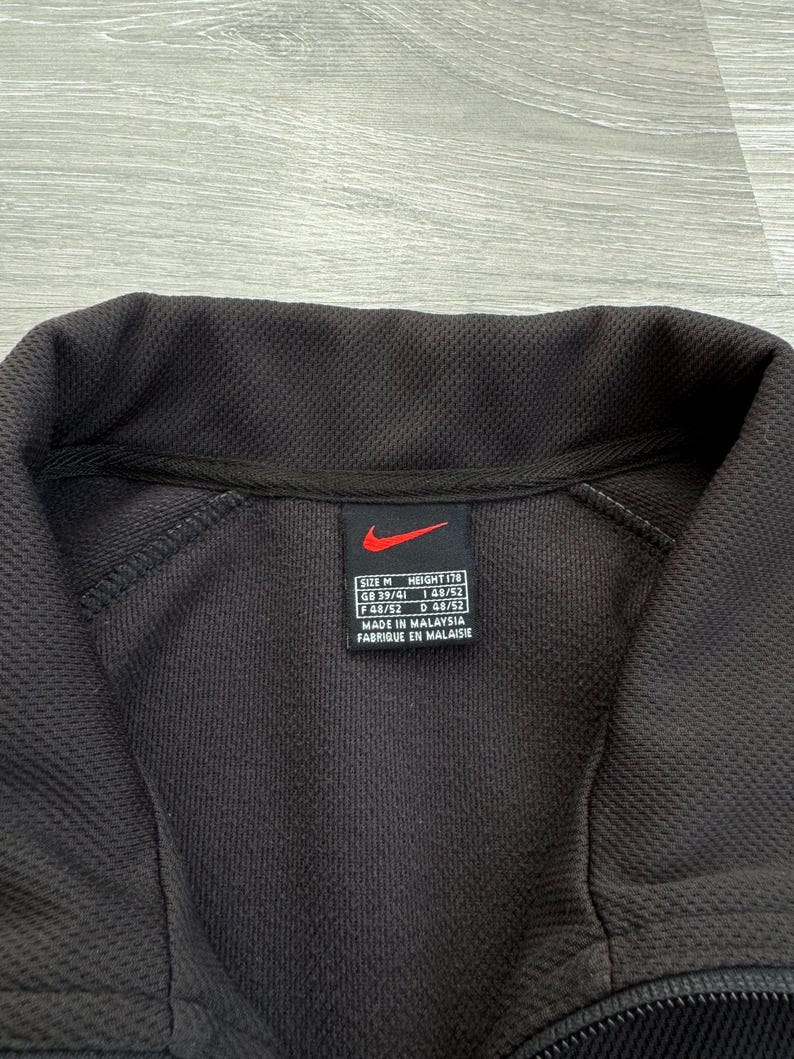 Puede incluir: Primer plano de una chaqueta deportiva negra Nike. La chaqueta tiene un patr&oacute;n texturizado y un cuello negro. Se ve una etiqueta con el logotipo de Nike e informaci&oacute;n sobre la talla. La chaqueta est&aacute; hecha en Malasia.