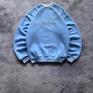 以下が含まれることがあります： 胸元にカレッジフォントでNIKEの文字が刺繍され、その下に小さなナイキのスウッシュロゴが刺繍されたライトブルーのクルーネックトレーナー。長袖で、袖口と裾はリブ編みになっています。