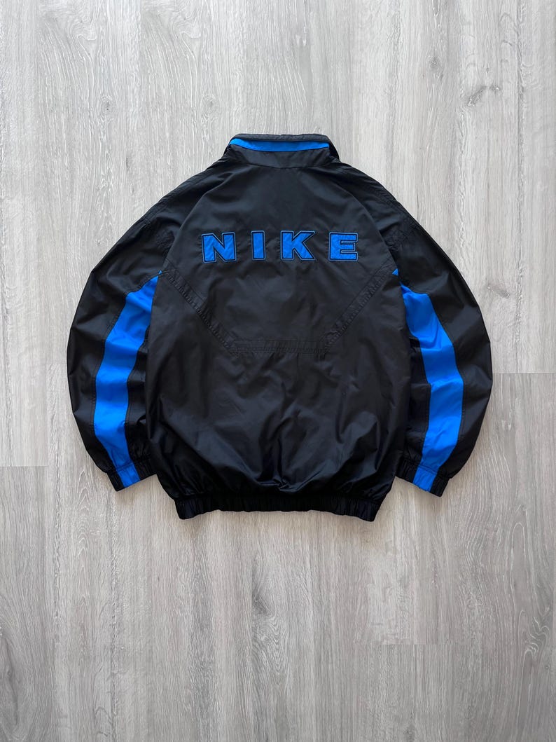 May include: Black Nike jacket with blue accents. The jacket features a blue Nike logo across the back and blue stripes down the sleeves. The jacket has a collar and a zipper closure.