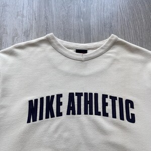 Puede incluir: Sudadera de color crema con el texto "NIKE ATHLETIC" en azul marino en el pecho. La sudadera tiene un cuello acanalado y un dise&ntilde;o cl&aacute;sico de cuello redondo.