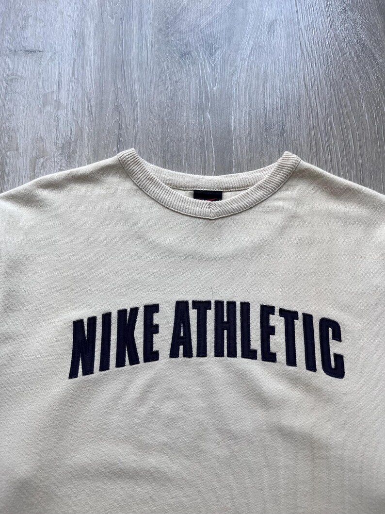 Puede incluir: Sudadera de color crema con el texto "NIKE ATHLETIC" en azul marino en el pecho. La sudadera tiene un cuello acanalado y un dise&ntilde;o cl&aacute;sico de cuello redondo.
