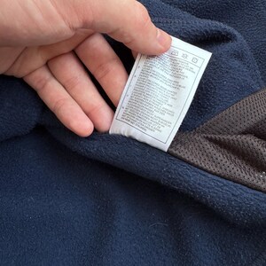 May include: Close-up of a navy blue fleece garment with a white tag. The tag has care instructions and text in multiple languages. A brown mesh material is visible on the right side of the image.