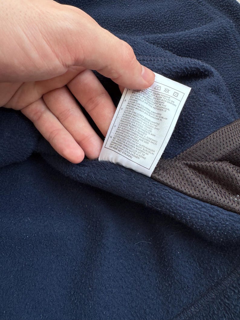May include: Close-up of a navy blue fleece garment with a white tag. The tag has care instructions and text in multiple languages. A brown mesh material is visible on the right side of the image.