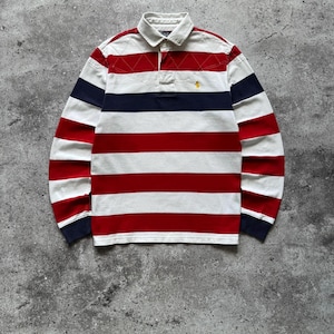 May include: A long-sleeve rugby shirt featuring horizontal stripes in red, white, and navy blue. The shirt has a white collar, a buttoned placket, and a small yellow embroidered logo on the left chest. The upper chest area includes a quilted diamond pattern.