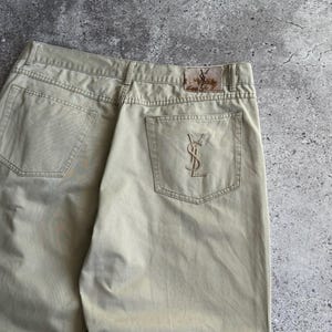 May include: Beige cotton trousers featuring a back pocket with an embroidered YSL logo in a matching thread colour. A rectangular leather-like patch is stitched onto the waistband, displaying the YSL logo in a dark, weathered brown finish.