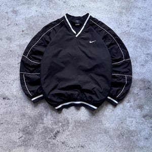 May include: A black long-sleeve pullover windbreaker featuring a V-neck collar with white trim, a small white Nike swoosh logo on the upper left chest, and white piping details along the sleeves and cuffs, finished with a white-striped hem.