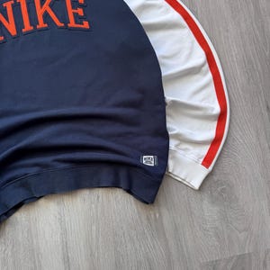 May include: Navy blue sweatshirt with the word "NIKE" in orange lettering across the chest. The right sleeve is white with a red stripe. A small Nike tag is on the lower left side.