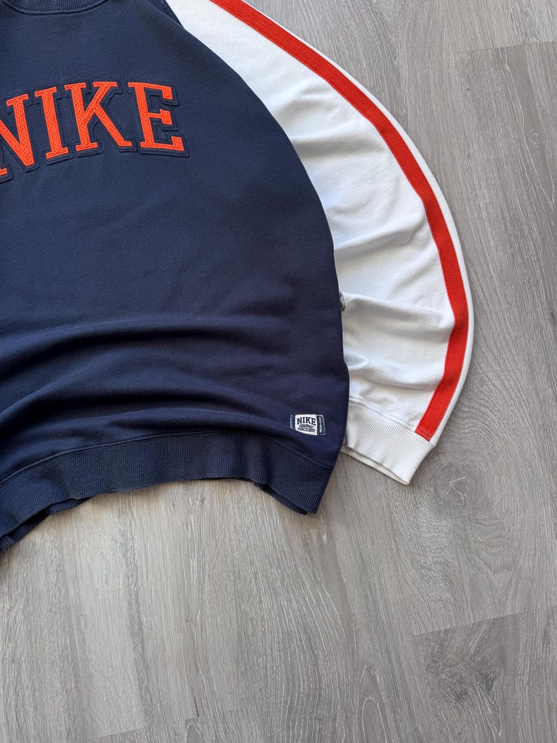 May include: Navy blue sweatshirt with the word "NIKE" in orange lettering across the chest. The right sleeve is white with a red stripe. A small Nike tag is on the lower left side.
