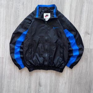 May include: Black Nike windbreaker jacket with blue accents. The jacket features a full-length zipper, a blue collar, and blue stripes down the sleeves. A small Nike logo is visible on the chest.