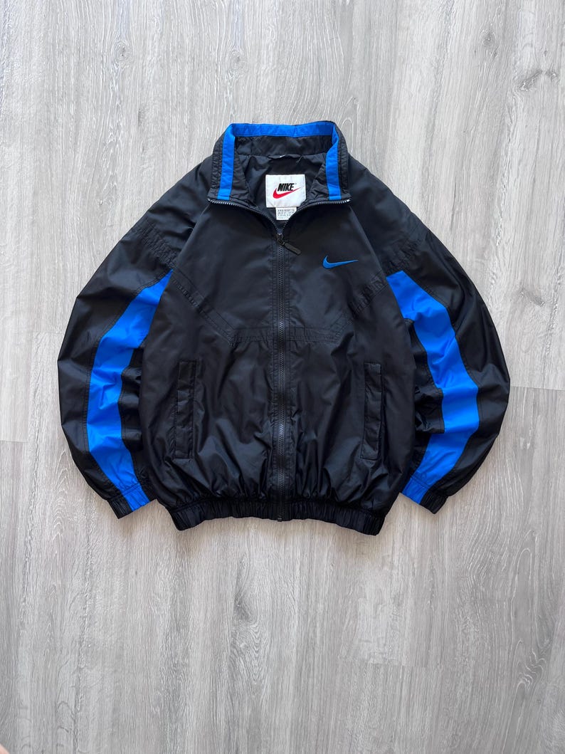 May include: Black Nike windbreaker jacket with blue accents. The jacket features a full-length zipper, a blue collar, and blue stripes down the sleeves. A small Nike logo is visible on the chest.