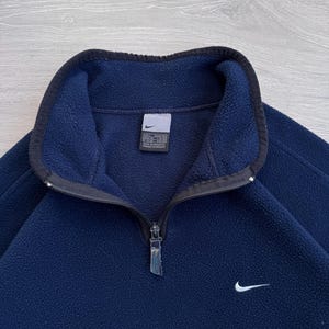 May include: Navy blue fleece pullover with a black collar and a quarter-zip closure. Features a small white Nike swoosh logo on the chest. The garment has a tag on the inside collar.
