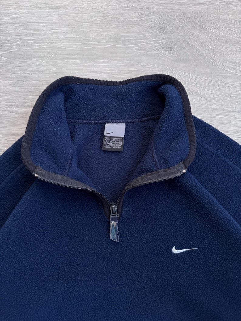 May include: Navy blue fleece pullover with a black collar and a quarter-zip closure. Features a small white Nike swoosh logo on the chest. The garment has a tag on the inside collar.