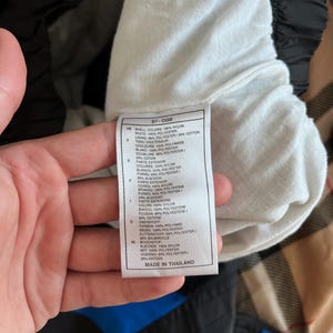 May include: A close-up of a tag with text, held by a hand. The tag lists fabric composition in multiple languages, including nylon, polyester, and cotton. The tag reads "MADE IN THAILAND".