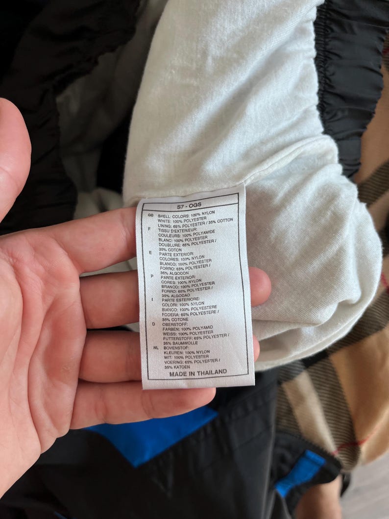 May include: A close-up of a tag with text, held by a hand. The tag lists fabric composition in multiple languages, including nylon, polyester, and cotton. The tag reads "MADE IN THAILAND".