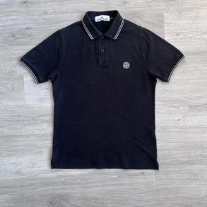 Pode incluir: Polo preto com detalhes brancos na gola e mangas. Possui uma carcela de dois botões e um pequeno patch com o logotipo no peito. A camisa é feita de um tecido macio e respirável.