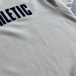Puede incluir: Una sudadera de color crema con la palabra "ATHLETIC" bordada en azul marino en el pecho. La tela parece suave y ligeramente texturizada. La imagen es un primer plano de la parte superior de la prenda.