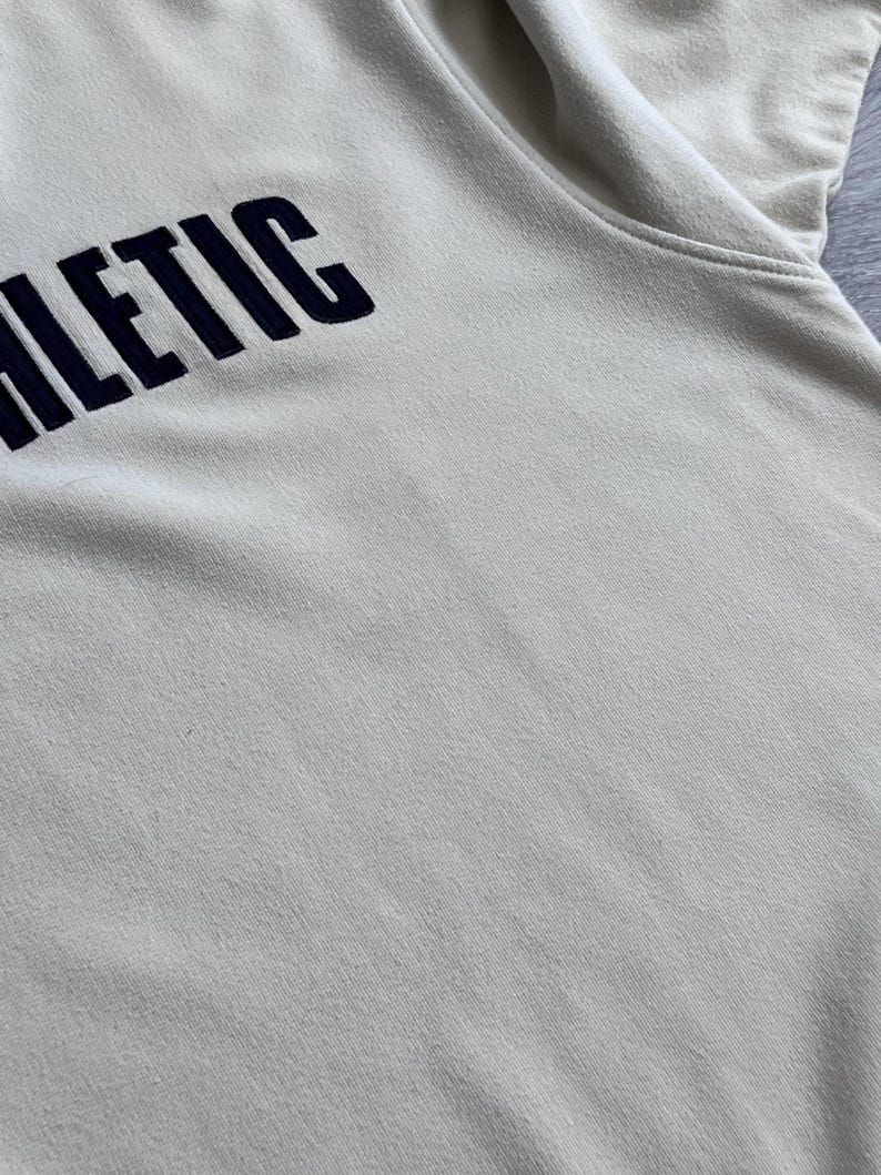 Puede incluir: Una sudadera de color crema con la palabra "ATHLETIC" bordada en azul marino en el pecho. La tela parece suave y ligeramente texturizada. La imagen es un primer plano de la parte superior de la prenda.