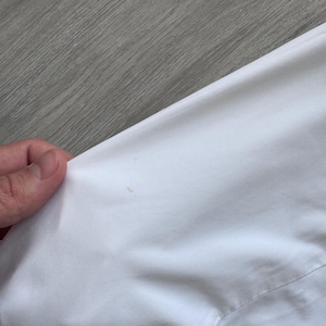 Peut inclure: Gros plan sur un v&ecirc;tement blanc, possiblement une chemise ou une veste, avec une couture visible et une petite tache. Le tissu semble lisse et propre, avec un l&eacute;ger &eacute;clat. L'image est prise en biais, montrant la texture du mat&eacute;riau.