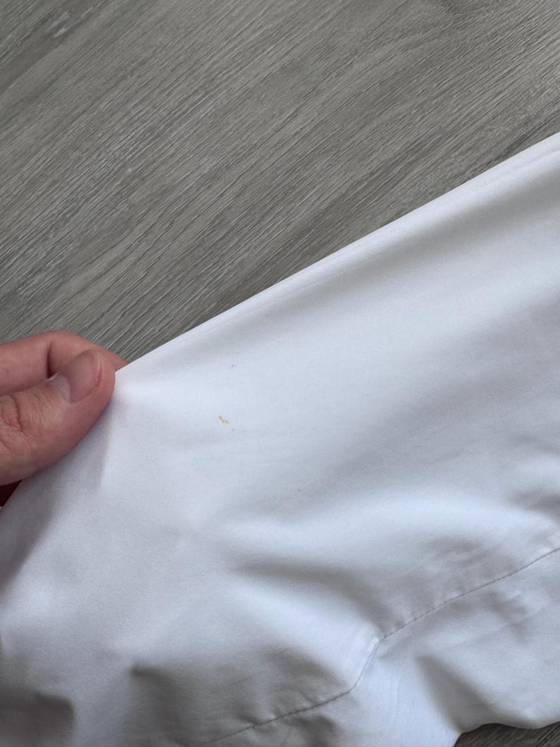 Peut inclure: Gros plan sur un v&ecirc;tement blanc, possiblement une chemise ou une veste, avec une couture visible et une petite tache. Le tissu semble lisse et propre, avec un l&eacute;ger &eacute;clat. L'image est prise en biais, montrant la texture du mat&eacute;riau.