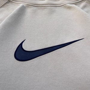 May include: A beige crew neck sweatshirt with a navy blue embroidered Nike swoosh logo. The sweatshirt has a ribbed collar and cuffs. The fabric appears to be a soft, comfortable material.