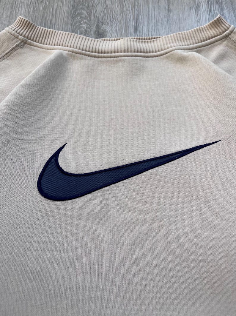 May include: A beige crew neck sweatshirt with a navy blue embroidered Nike swoosh logo. The sweatshirt has a ribbed collar and cuffs. The fabric appears to be a soft, comfortable material.