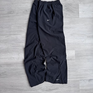 Puede incluir: Pantalones deportivos negros con un logotipo blanco de Nike. Los pantalones tienen cintura el&aacute;stica y corte holgado. Est&aacute;n hechos de un material ligero, posiblemente nailon. Tienen una abertura en la parte inferior de cada pierna.