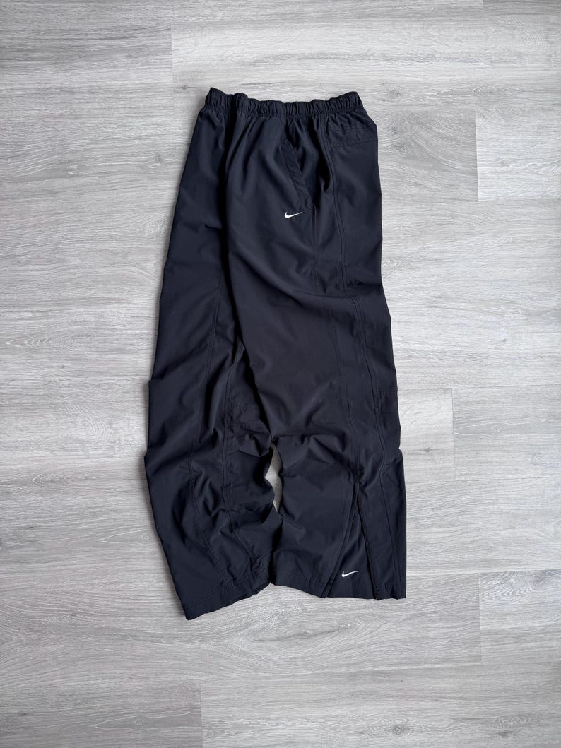 Puede incluir: Pantalones deportivos negros con un logotipo blanco de Nike. Los pantalones tienen cintura el&aacute;stica y corte holgado. Est&aacute;n hechos de un material ligero, posiblemente nailon. Tienen una abertura en la parte inferior de cada pierna.