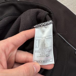 Puede incluir: Primer plano de una chaqueta deportiva negra con una etiqueta blanca. La etiqueta dice "MADE IN MALAYSIA" y enumera la composici&oacute;n de la tela como 100% poli&eacute;ster. La chaqueta tiene un tejido texturizado y una raya plateada reflectante.