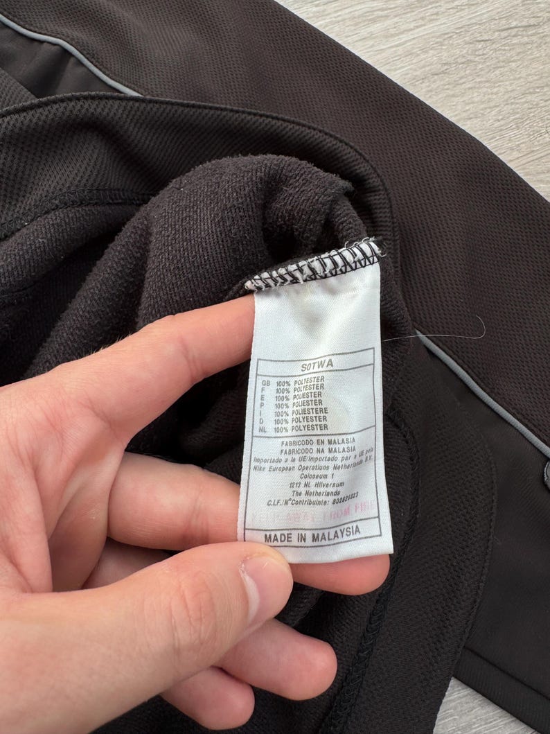 Puede incluir: Primer plano de una chaqueta deportiva negra con una etiqueta blanca. La etiqueta dice "MADE IN MALAYSIA" y enumera la composici&oacute;n de la tela como 100% poli&eacute;ster. La chaqueta tiene un tejido texturizado y una raya plateada reflectante.