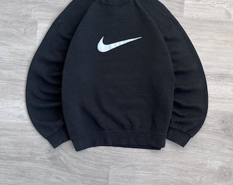 Puede incluir: Sudadera negra de cuello redondo con el logotipo blanco de Nike en la parte delantera. La parte superior de manga larga está hecha de un material suave y está colocada sobre una superficie de madera.