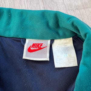 May include: Close-up of a navy blue and teal Nike track jacket collar. The Nike logo is red on a white tag. The jacket's tag includes washing instructions and size information.