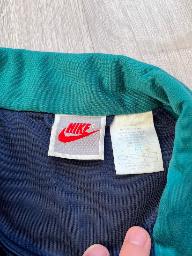 May include: Close-up of a navy blue and teal Nike track jacket collar. The Nike logo is red on a white tag. The jacket's tag includes washing instructions and size information.