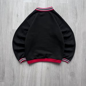 May include: Black sweatshirt with a red and white striped collar and cuffs. The crew neck sweatshirt has long sleeves. The bottom hem is red with white stripes. The garment is displayed on a light grey wooden surface.