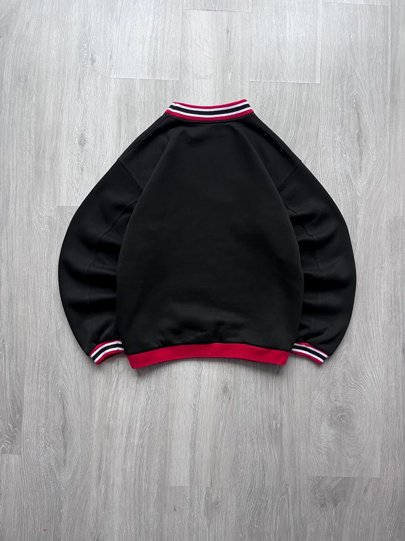 May include: Black sweatshirt with a red and white striped collar and cuffs. The crew neck sweatshirt has long sleeves. The bottom hem is red with white stripes. The garment is displayed on a light grey wooden surface.