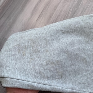 May include: Close-up of a light gray fabric with brown stains. The fabric appears to be a section of a garment, possibly a sleeve or a similar part. The fabric is slightly wrinkled.
