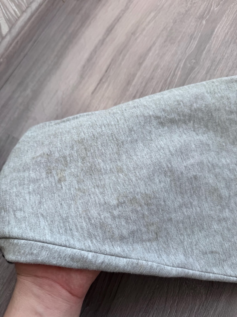 May include: Close-up of a light gray fabric with brown stains. The fabric appears to be a section of a garment, possibly a sleeve or a similar part. The fabric is slightly wrinkled.