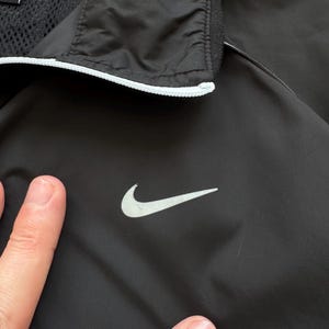 Peut inclure: Veste de sport noire avec le logo Nike blanc sur la poitrine. La veste a une bordure blanche autour du col et une doublure en mesh. La veste est faite d'un mat&eacute;riau l&eacute;ger.