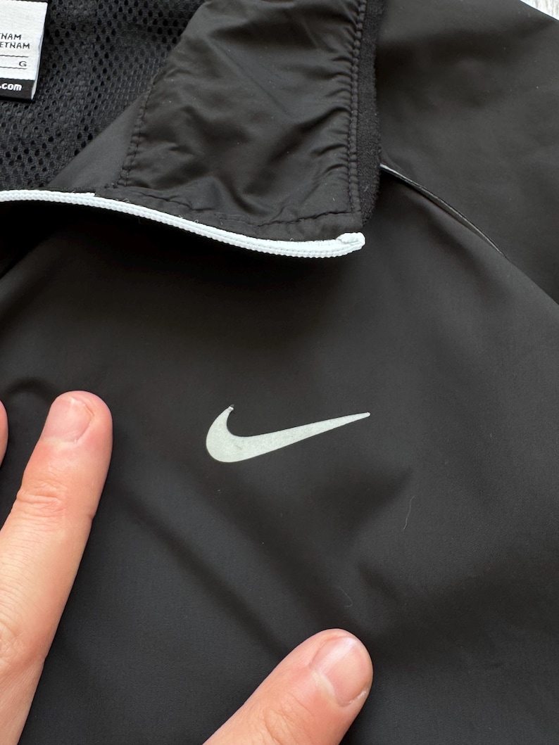Peut inclure: Veste de sport noire avec le logo Nike blanc sur la poitrine. La veste a une bordure blanche autour du col et une doublure en mesh. La veste est faite d'un mat&eacute;riau l&eacute;ger.