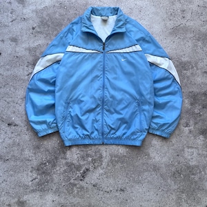 May include: A light blue zip-up track jacket featuring a white panel across the chest and shoulders with thin black piping. The jacket has a high collar, long sleeves with elastic cuffs, two zippered side pockets, and a small white Nike logo.